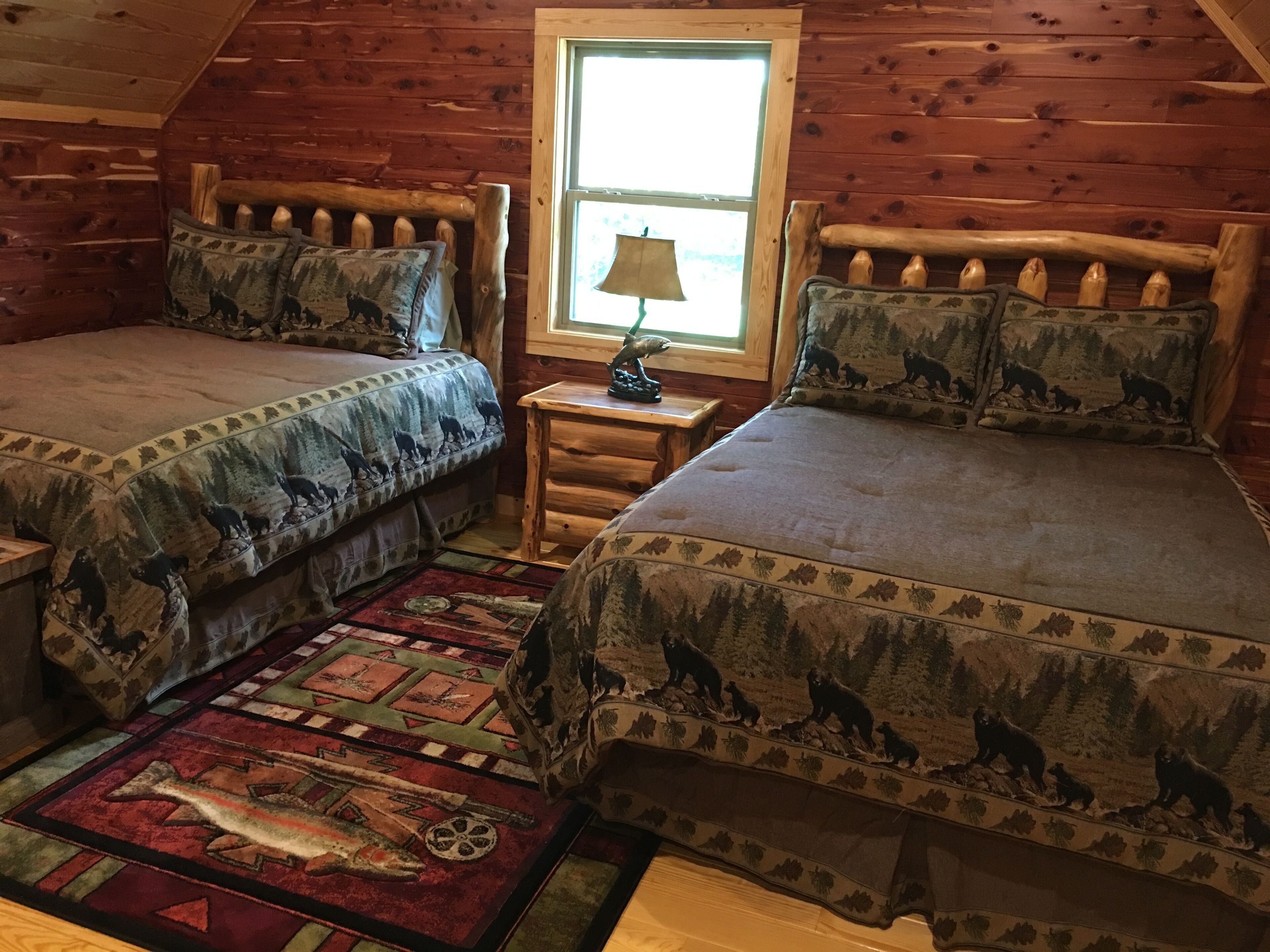 A cozy cabin room with two wooden beds, a window with a view, and a small table with a lamp.