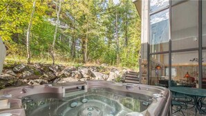 Un bain à remous installé dans un cadre naturel, avec vue sur une forêt et un bâtiment doté de grandes fenêtres.