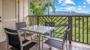 Un balcon avec une table et des chaises, donnant sur un jardin luxuriant et offrant une vue panoramique sur l’océan.