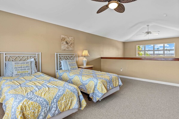 A bedroom with two beds, a ceiling fan, a nightstand with a lamp, a window with a view, and a decorative wall piece.