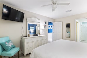 Une chambre à coucher comprenant un lit, une commode, un téléviseur fixé au mur, un ventilateur de plafond et une porte donnant sur une autre pièce.