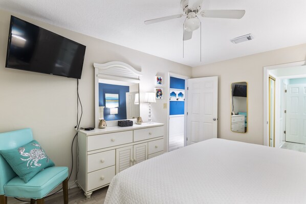 Une chambre à coucher comprenant un lit, une commode, un téléviseur fixé au mur, un ventilateur de plafond et une porte donnant sur une autre pièce.