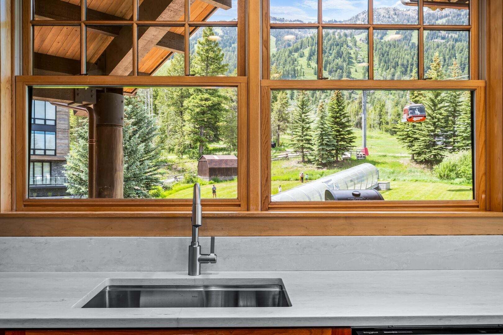 A kitchen with a stainless steel sink and wooden window frames overlooking a mountainous landscape.