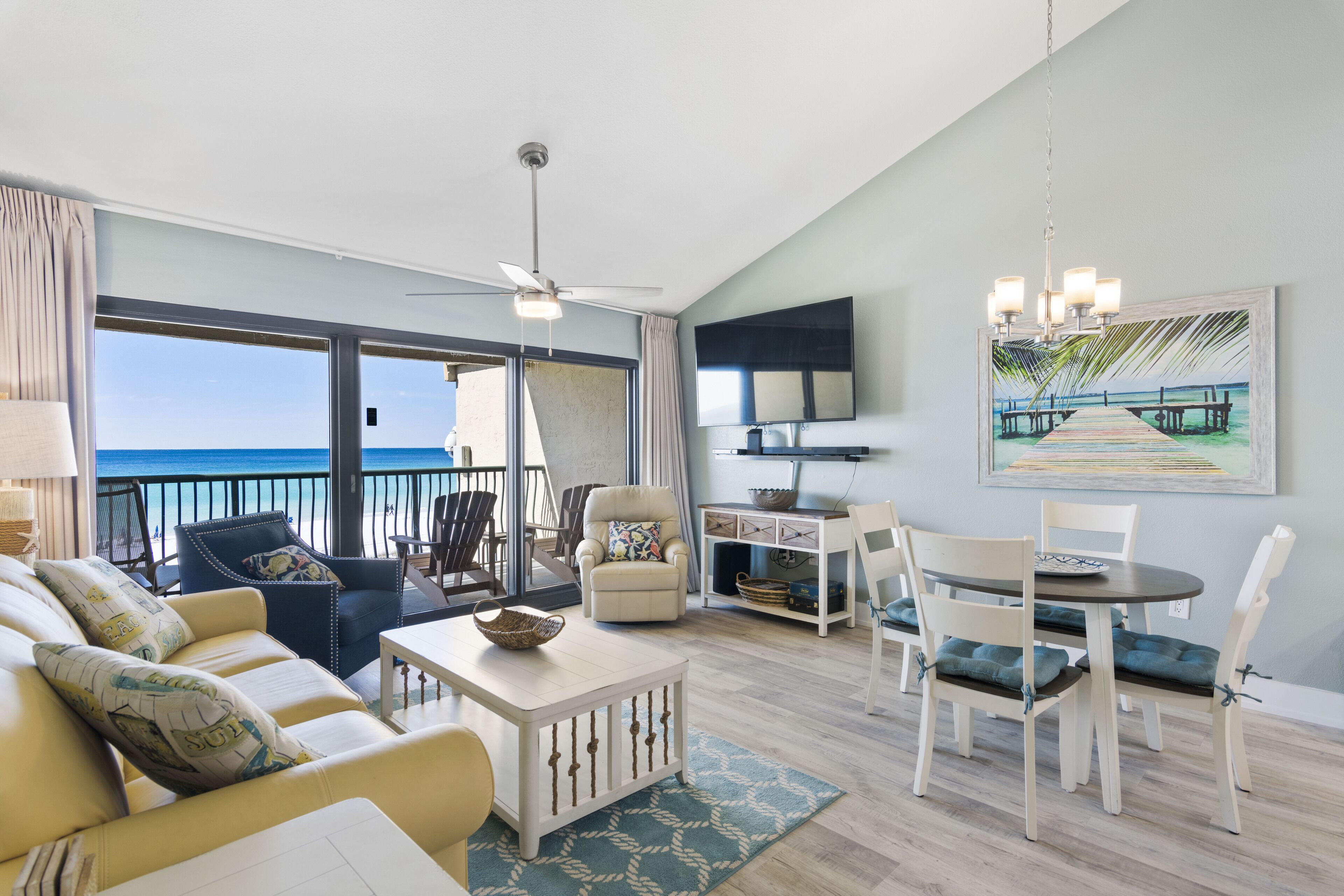 A spacious living room with a balcony overlooking the ocean, a dining area, and a TV mounted on the wall.