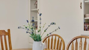 Une table à manger surmontée d’un pichet blanc rempli de fleurs bleues, entourée de chaises en bois.