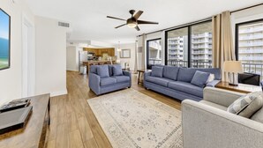 Un salon avec deux canapés, une table basse et un téléviseur. On y trouve un ventilateur de plafond, une grande fenêtre donnant sur un immeuble, et une cuisine à l’arrière-plan.