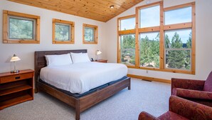 Ein Schlafzimmer mit einem hölzernen Bett, zwei Nachttischen, einem Sessel und großen Fenstern mit Blick auf Bäume.