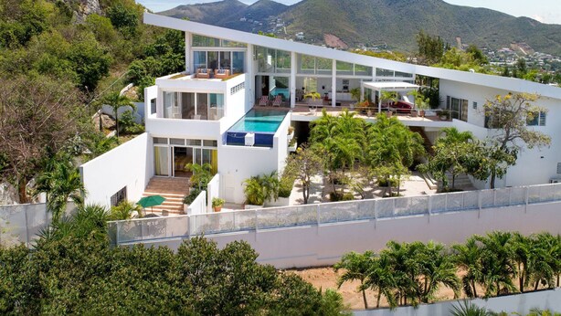 A modern multi-story house with a pool, surrounded by lush greenery and mountains.