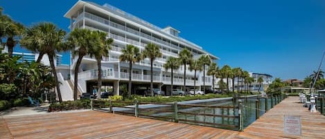 Un hotel moderno con varios balcones, un paseo marítimo de madera y palmeras.