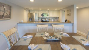 A dining area with a wooden table set for four, a kitchen in the background, and a starfish artwork on the wall. - Beautiful San Remo on Beach, Water-view From All Windows, Heated Pool, Hot Tub  (Redington Shores)