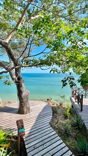 Een houten terras dat naar een strand leidt, met helder turquoise water, omringd door weelderige begroeiing en een grote boom.