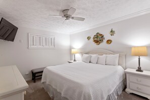 Une chambre à coucher avec un grand lit, deux tables de chevet avec des lampes, un ventilateur de plafond, un banc et des œuvres d’art décoratives accrochées au mur.