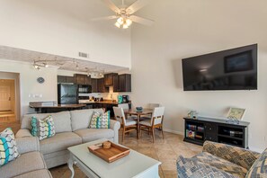 A spacious living area with a sofa, coffee table, and dining table. A kitchen is visible in the background, and a flat-screen TV is mounted on the wall.