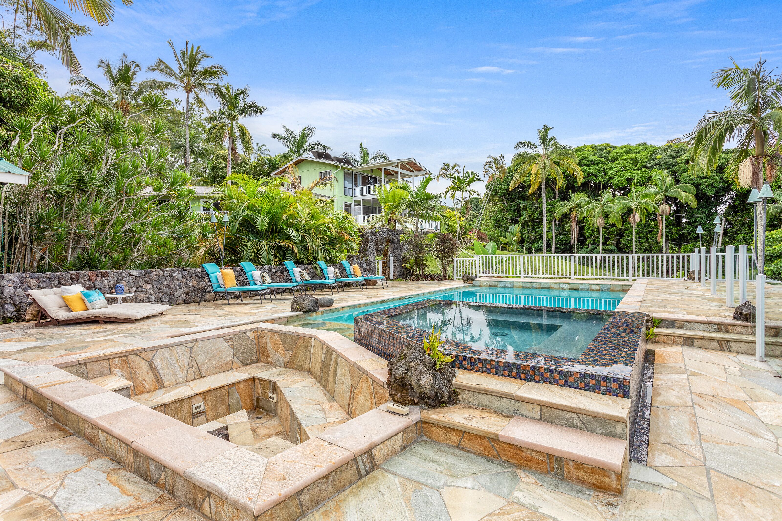 A pool area with stone steps, lounge chairs, and a view of a house surrounded by palm trees.