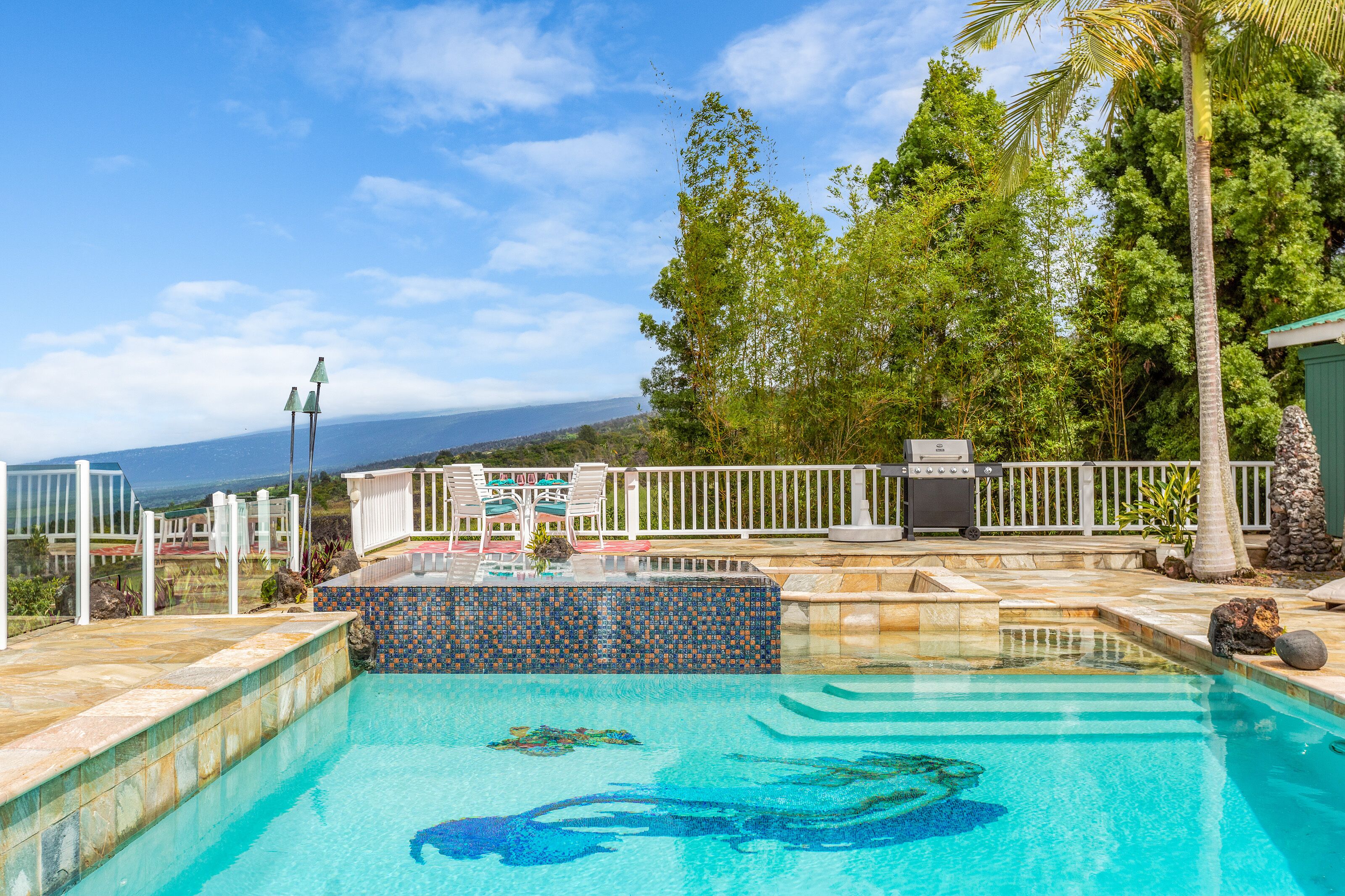 A swimming pool with a tiled edge, a lounge area with chairs, a grill, and a view of the ocean.