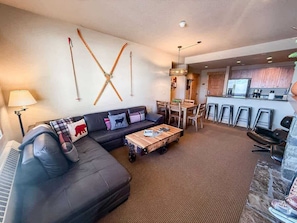 A modern living room with a sofa, coffee table, and a dining area with chairs and a bar. - Ski In / Ski Out Condo, Amazing Views, Sleeps 8! (Whitefish)