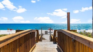 Une terrasse en bois mène à une plage où l’eau est d’un bleu cristallin, et où l’on aperçoit quelques personnes au loin.