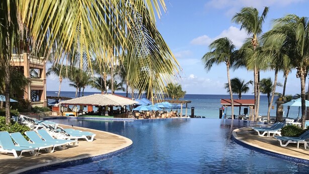 A pool area with lounge chairs and umbrellas, surrounded by palm trees and a view of the ocean. - “Our Paradise Found” Condo 1405 at Infinity Bay (West Bay Beach, Roatan)