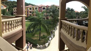 A view from a balcony overlooking a courtyard with palm trees and a building with red-tiled roofs. - “Our Paradise Found” Condo 1405 at Infinity Bay (West Bay Beach, Roatan)