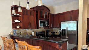 A kitchen with dark wooden cabinets, a stainless steel refrigerator, a microwave, and a coffee maker. There are two wooden chairs with cushioned seats. - “Our Paradise Found” Condo 1405 at Infinity Bay (West Bay Beach, Roatan)