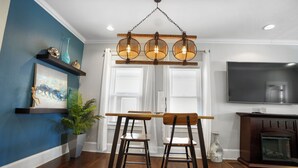 Un comedor con una mesa de madera y dos taburetes altos, una lámpara colgante, un televisor y una chimenea.