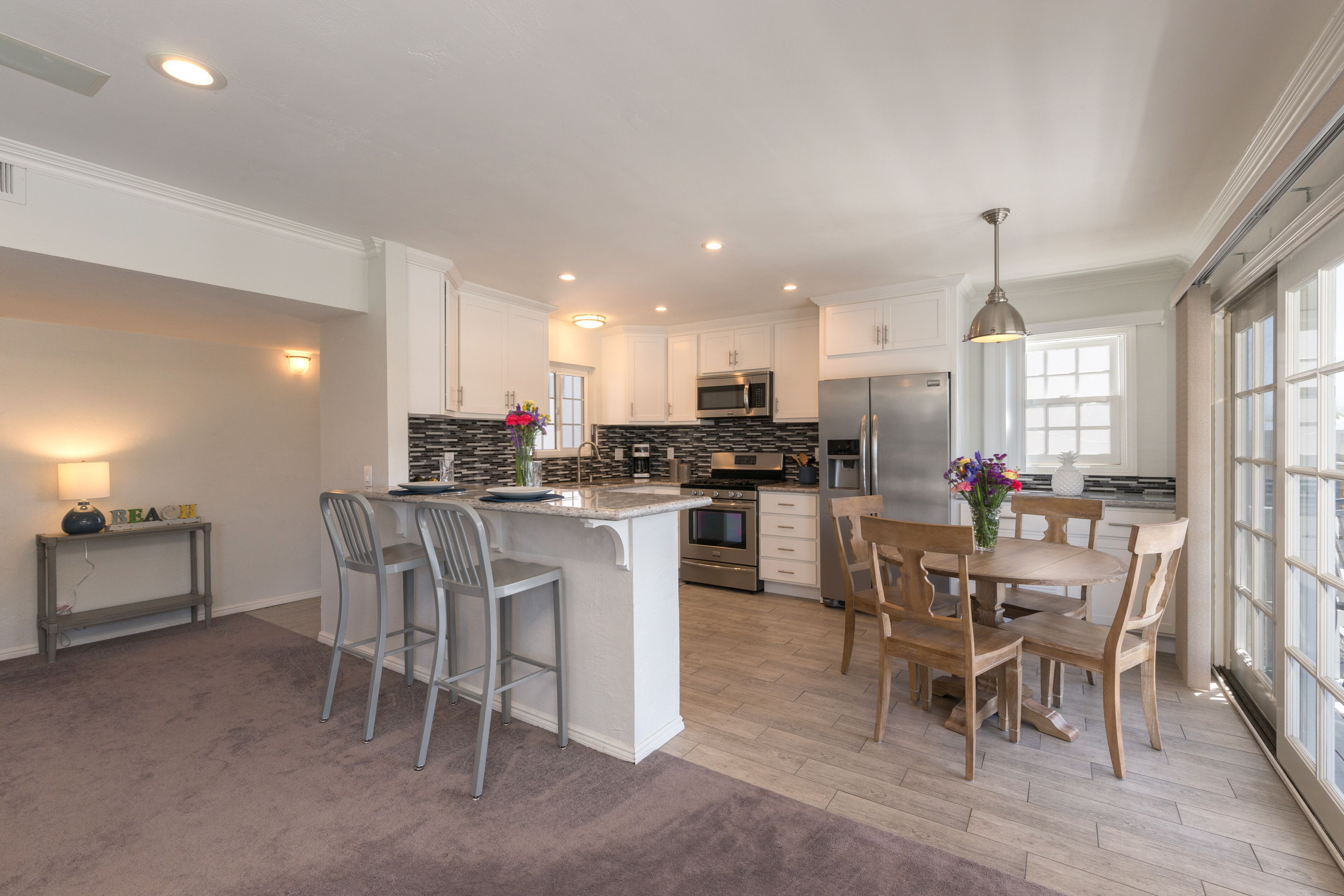 A modern kitchen with white cabinets, a central island with bar stools, a dining table with chairs, and a large window providing natural light.