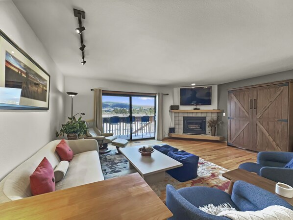 A spacious living room with a sofa, coffee table, and blue chairs. A fireplace and TV are mounted on the wall.