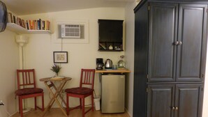 Ein kompakter Wohnbereich mit Küchenzeile, zwei Stühlen, einem kleinen Tisch, einem Kühlschrank, einem Schrank, einem Heizkörper und einem Bücherregal.