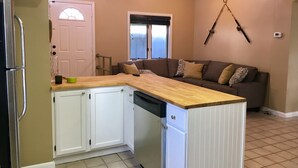 Una cocina compacta con armarios blancos, encimera de madera y lavavajillas. Un sofá marrón con cojines estampados está apoyado en la pared, y una caña de pescar está colgada en la pared.