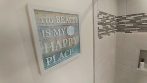 Een badkamer met een betegelde muur, een lijst met de tekst 'THE BEACH IS MY HAPPY PLACE' en een klein plankje met een fles.