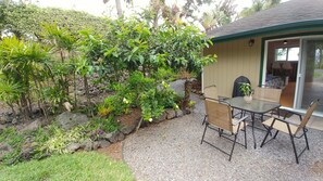 Un espace patio aménagé avec une table et des chaises, entouré d’un jardin où poussent différentes plantes et agrémenté d’un petit jardin de rocaille.