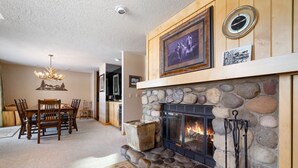 A cozy living room with a stone fireplace, a dining area with wooden furniture, and a chandelier. - RMR: Greyling 4513 Condo in The Aspens/Wilson,WY (Wilson)