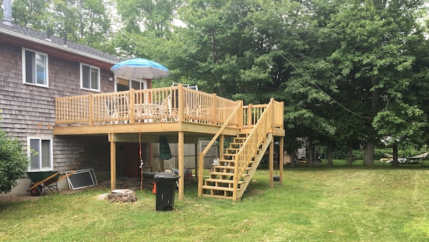 A wooden deck with a staircase leading to a covered area, surrounded by a grassy yard and trees.