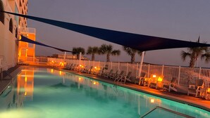 A swimming pool with lounge chairs and palm trees, surrounded by a white fence and a building with balconies.