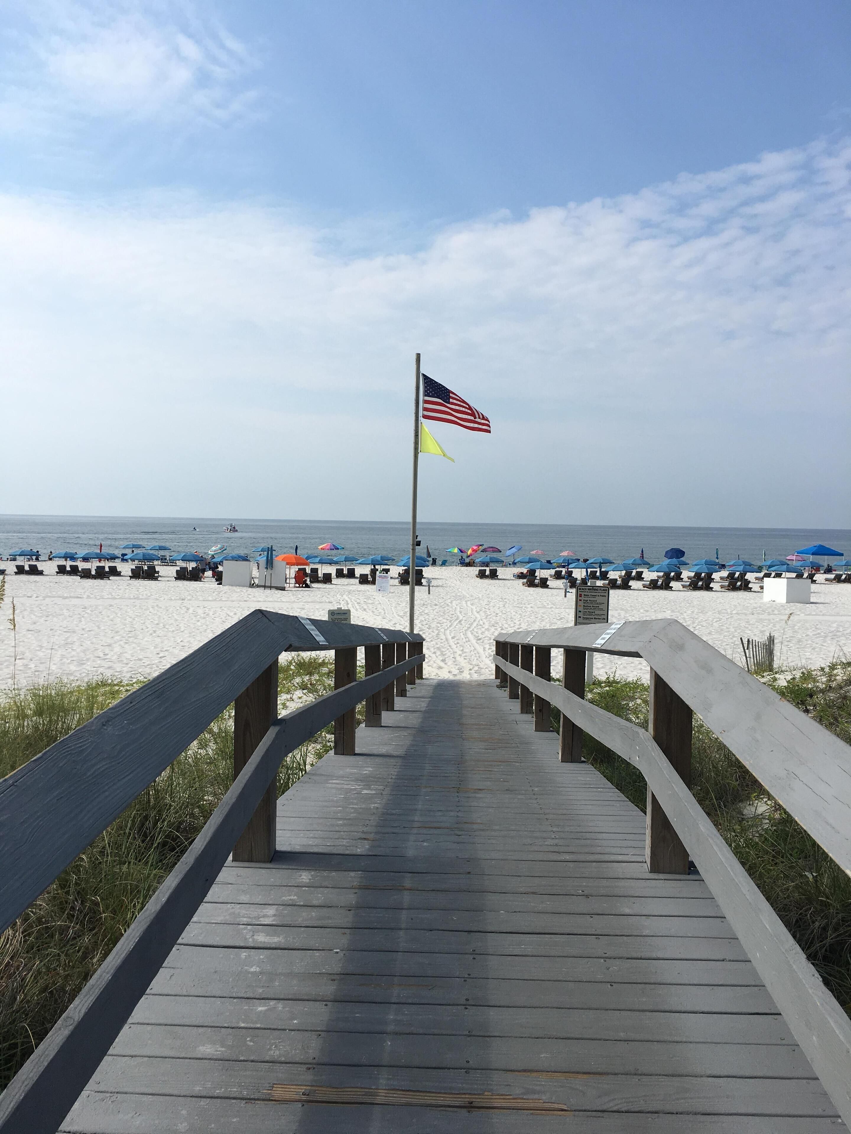 Une promenade en bois menant à une plage agrémentée de parasols et d’un drapeau.