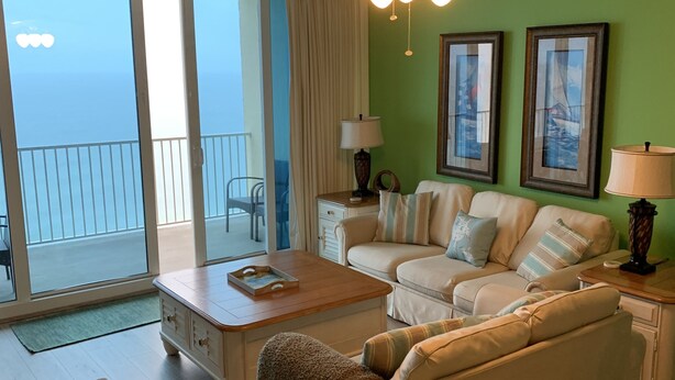 A living room with a sofa, a coffee table, and a ceiling fan. There are two lamps on side tables, and a window with curtains.