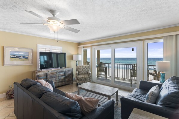 A living room with a sofa, coffee table, and a television. There is a balcony with a view of the ocean.