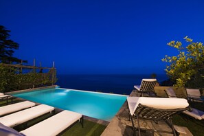An infinity pool with white lounge chairs, a wooden pergola, and a view of the ocean at dusk.
