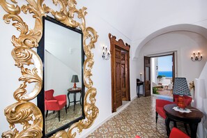 Un pasillo con un espejo grande y ornamentado, sillas rojas, un suelo estampado y vista al mar a través de una puerta abierta.