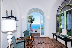 Una habitación con vista al mar, una silla, una mesa con un florero y una lámpara.