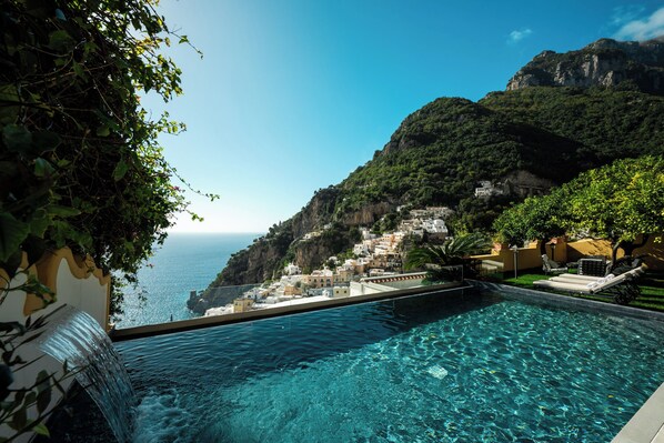 A pool with clear blue water, surrounded by lush greenery and overlooking a coastal village with white buildings.