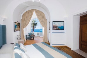 Un dormitorio con una cama grande, vista al mar a través de un ventanal arqueado y una puerta que da a un balcón.