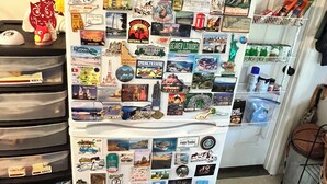 Un refrigerador cubierto de imanes y calcomanías, incluyendo objetos de viaje y una foto de manos sosteniendo a un bebé.