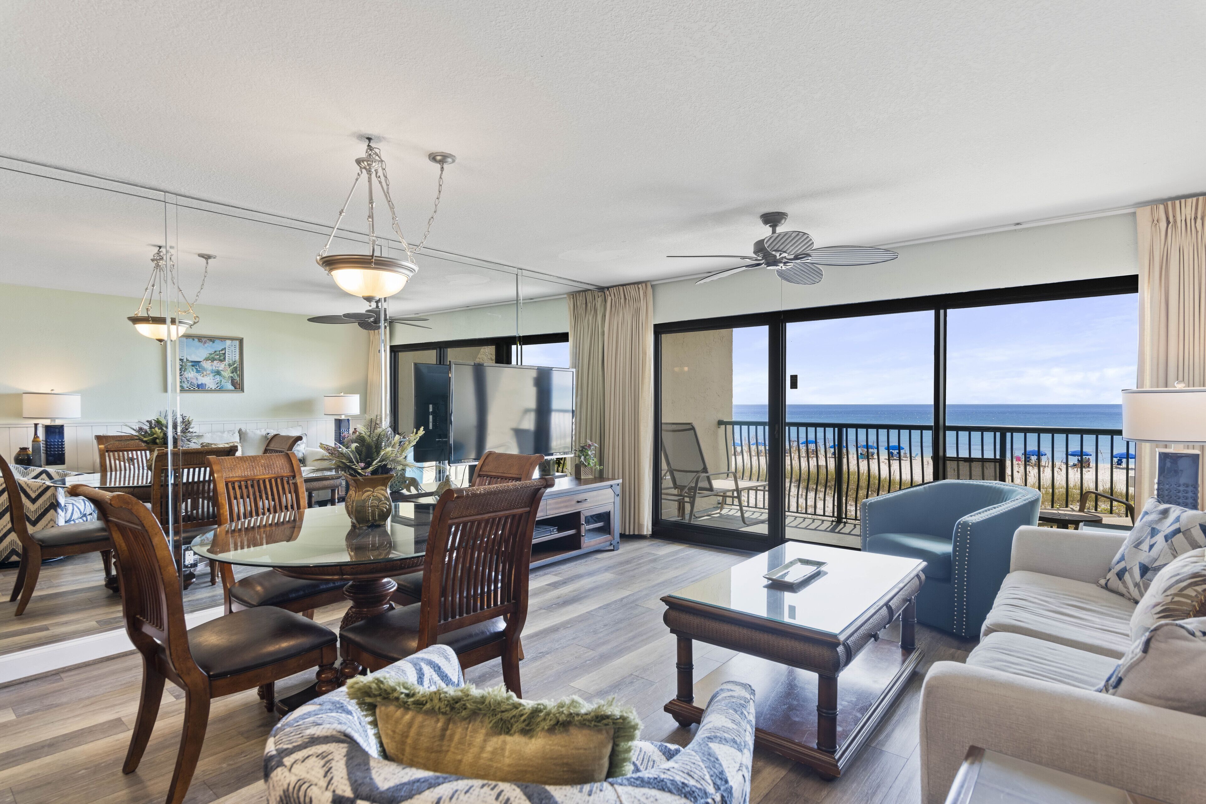 A spacious living area with a large glass door leading to a balcony with a view of the ocean. There is a dining table with chairs, a sofa, and a coffee table.