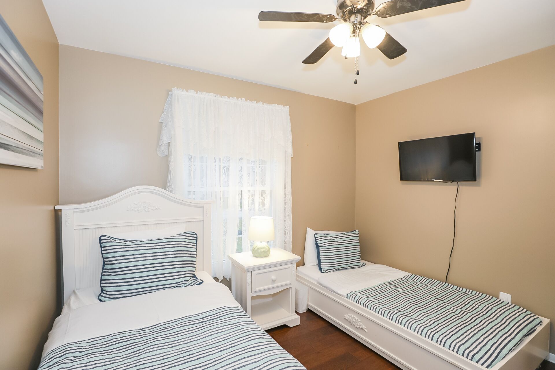 A bedroom with two single beds, a ceiling fan, a TV mounted on the wall, and a window with sheer curtains.