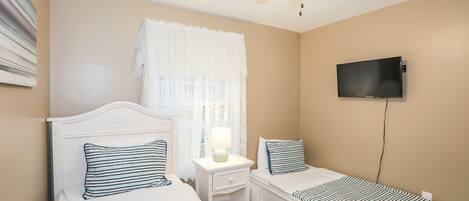 A bedroom with two single beds, a ceiling fan, a TV mounted on the wall, and a window with sheer curtains.