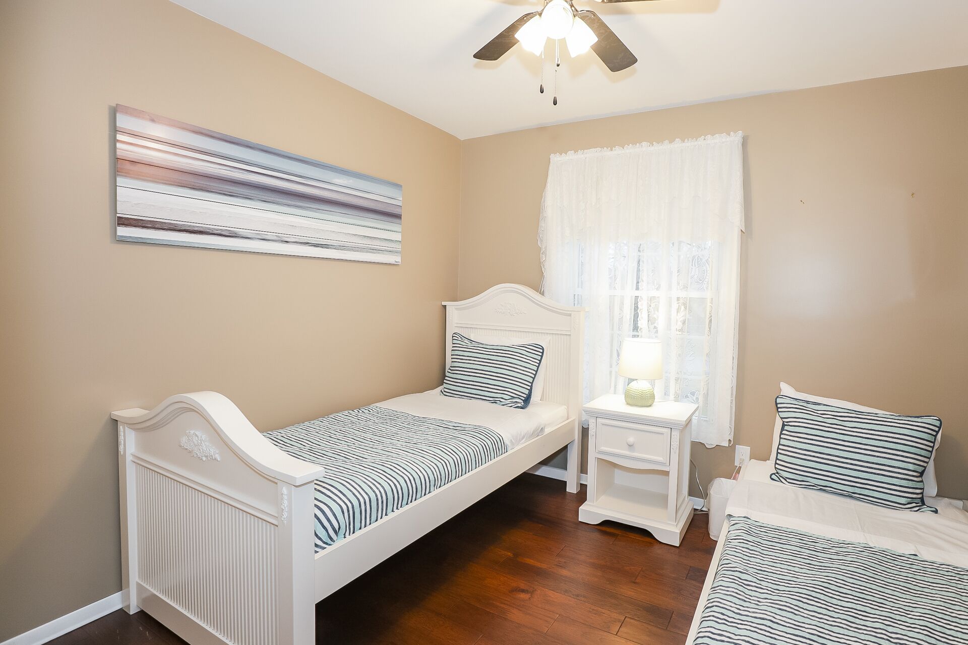 A bedroom with two single beds, a ceiling fan, a window with sheer curtains, and a wall-mounted abstract painting.