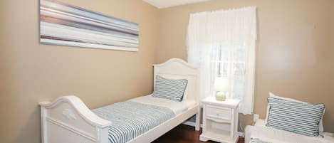 A bedroom with two single beds, a ceiling fan, a window with sheer curtains, and a wall-mounted abstract painting.