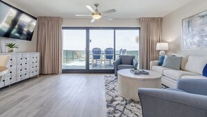 A modern living room with a flat-screen TV, a ceiling fan, a sofa, a coffee table, and a balcony with outdoor seating.