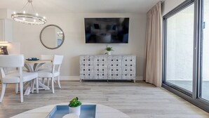 A modern living room with a dining area, a large mirror, a flat-screen TV, and a large window with curtains.